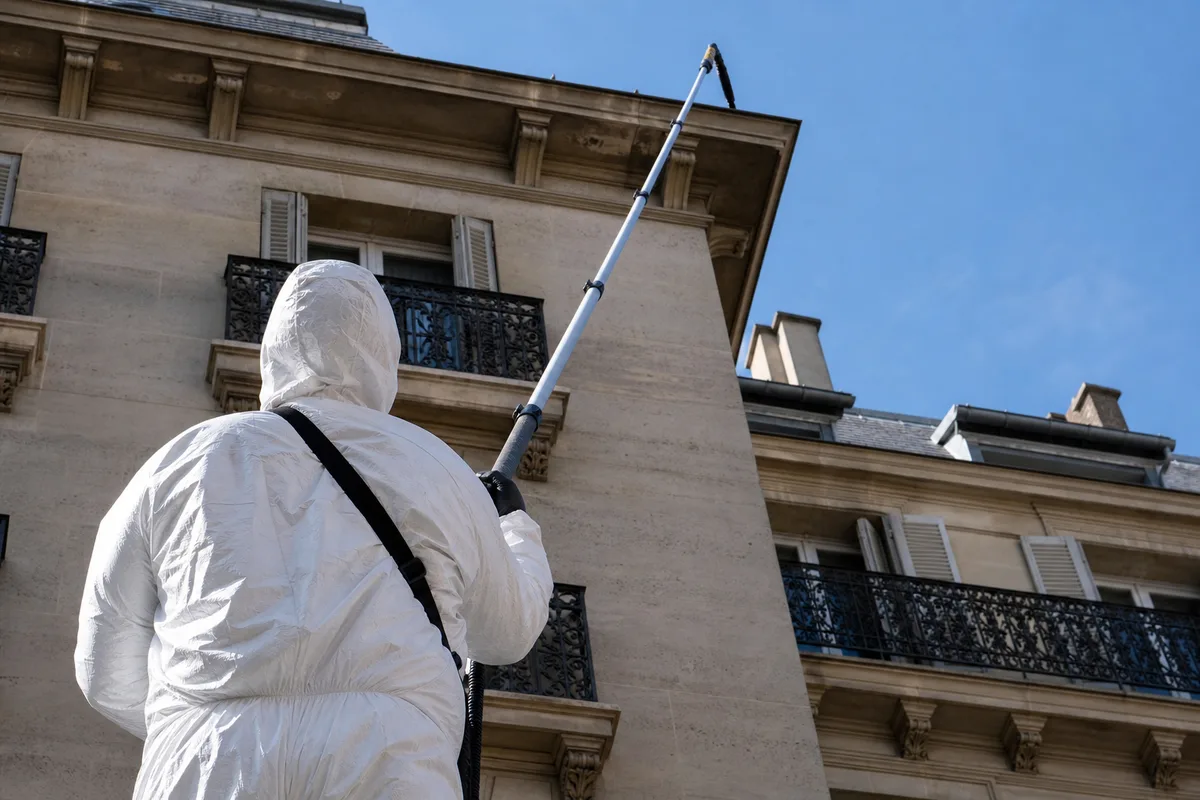 Intervenant en combinaison de protection utilisant une perche télescopique vers une toiture parisienne
